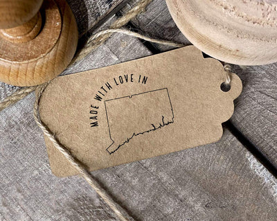 A craft tag stamped with the phrase Made with Love in and an outline of a Connecticut state silhouette tied with twine next to a wooden stamp on a rustic wooden background