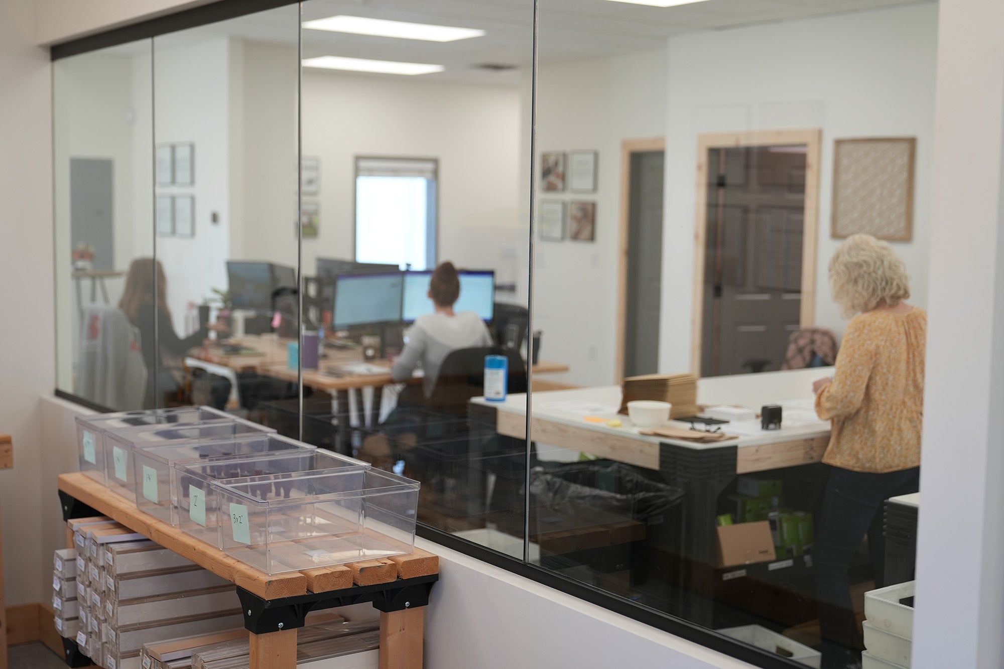 View through a glass partition of a bustling custom rubber stamp workshop with workers focused on their tasks crafting materials on desks and organized storage bins in the foreground