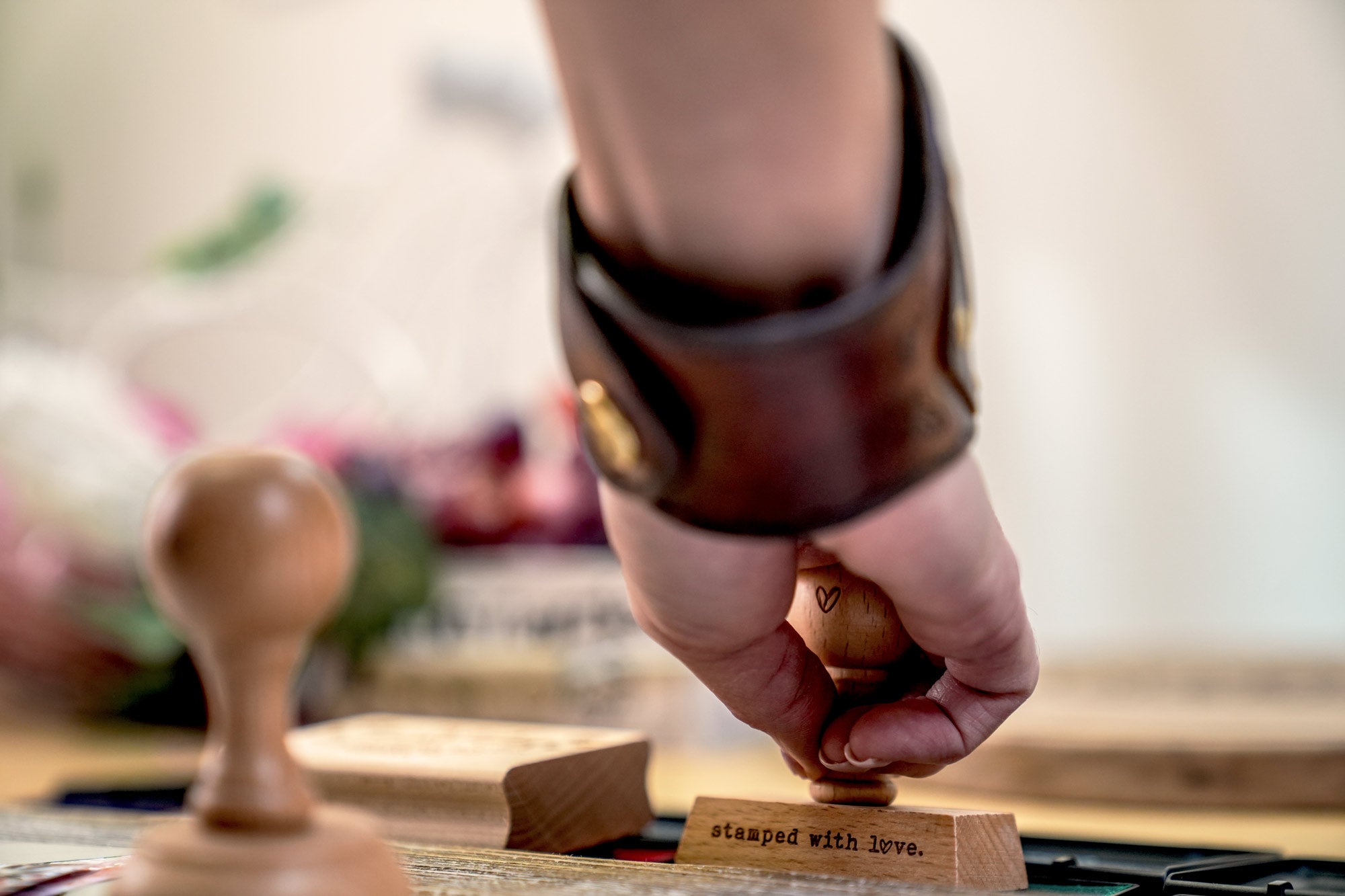 Close up of a hand holding a custom rubber stamp with the text stamped with love focusing on the stamping process with embossers and other craft tools softly blurred in the background
