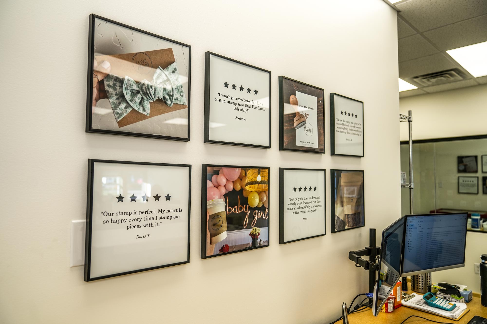 A wall display featuring framed customer testimonials and images of custom rubber stamp projects reflecting customer satisfaction and the personalized touch of the brand alongside a workstation with a computer monitor