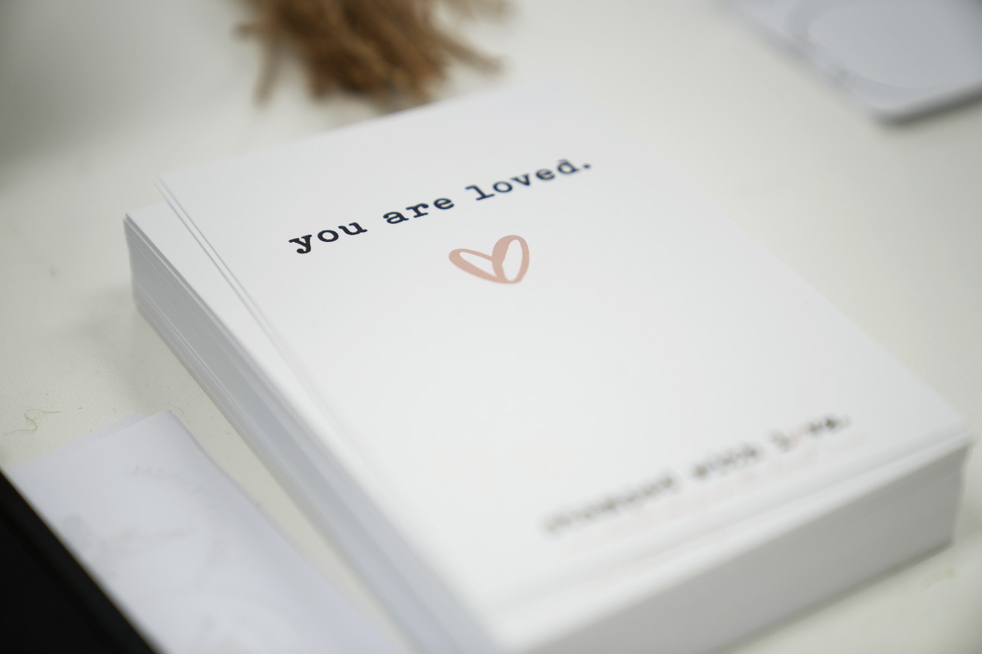 A stack of freshly printed cards with the phrase you are loved and a small pink heart showcasing the final product of the custom stamping process on a white worktable