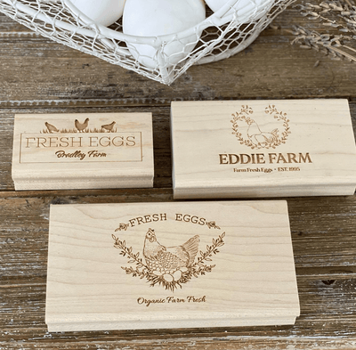  Three engraved wooden egg stamps featuring farm-themed designs and labels such as Fresh Eggs Bradley Farm and EDDIE FARM Farm Fresh Eggs and Fresh Eggs Organic Farm Fresh displayed on a rustic wooden surface with white eggs in a wire basket nearby.