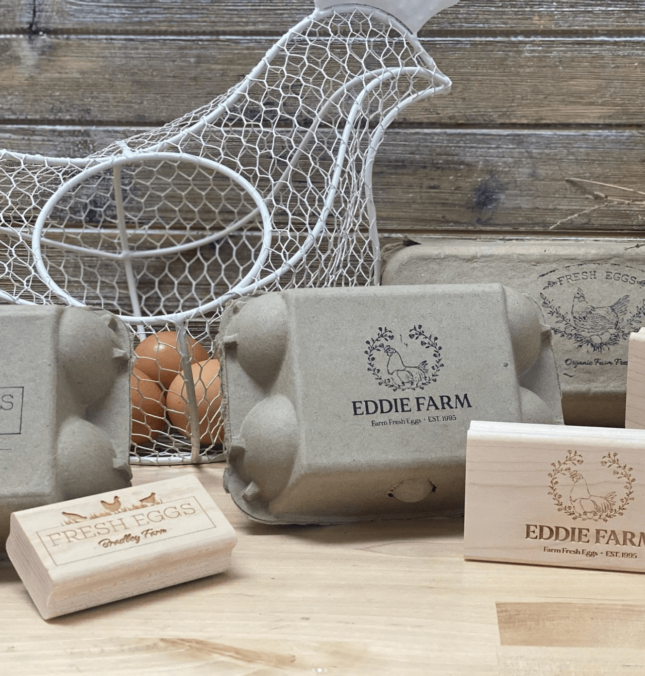 Rustic egg cartons and custom wood stamps labeled Eddie Farm and Fresh Eggs Bradley Farm displayed with a white wire chicken basket holding brown eggs, all set on a wooden surface with a weathered wood backdrop