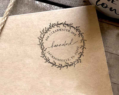 A return address stamp featuring an elegant circular leafy wreath design encircling a handwritten style with full address below