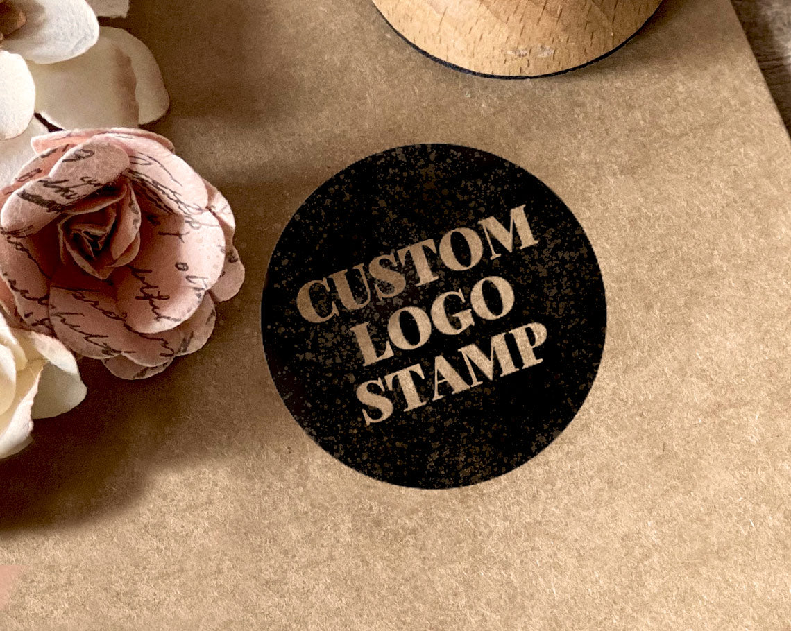 A circular black ink stamp on brown paper with the words CUSTOM LOGO STAMP beside paper flowers.