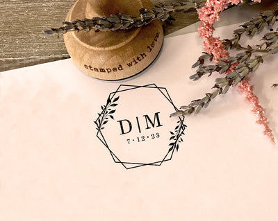 A hexagonal save the date stamp imprint with the initials D I M and a date framed by a leafy design on a paper surface next to a wooden stamp handle