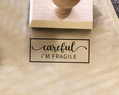 A paper bag stamped with the message careful I'm fragile beside a wooden rubber stamp handle on a fabric surface.