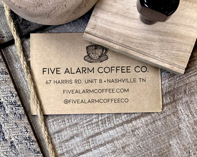 A business card for Five Alarm Coffee Co with address and social media details stamped on craft paper beside a rubber stamp and a spool of twine on a wooden background