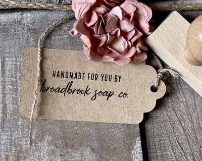 A craft tag with Handmade for you by Broadbrook Soap Co stamped on it tied with twine next to a wooden stamp and a pink fabric flower on a wooden surface