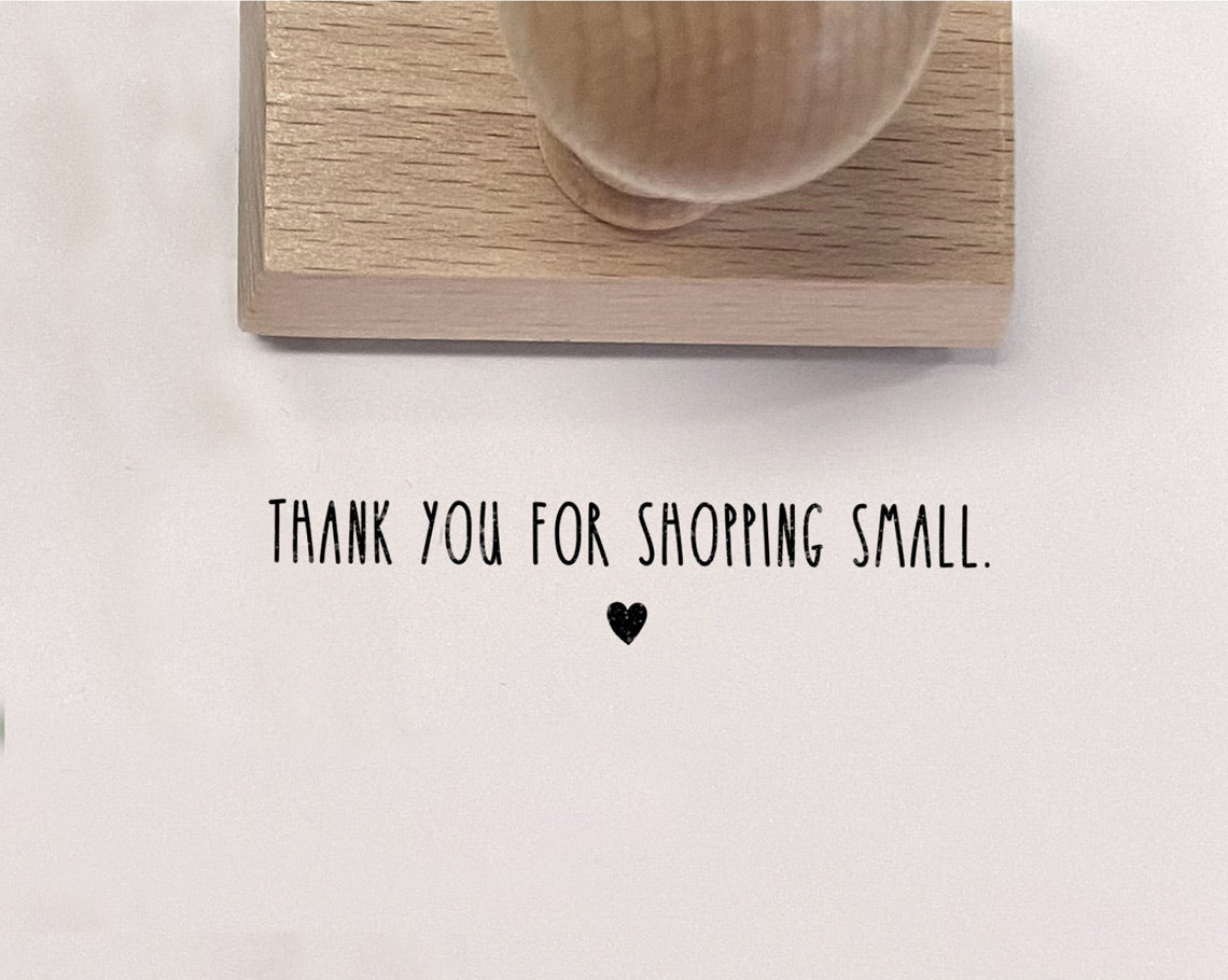 A piece of paper with the phrase Thank you for shopping small stamped on it next to a wooden rubber stamp with a heart symbol.