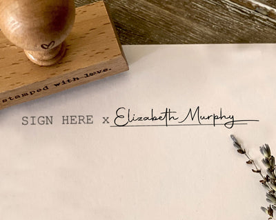 A rectangular rubber signature stamp with a full-name signature imprinted next to a sign here placeholder