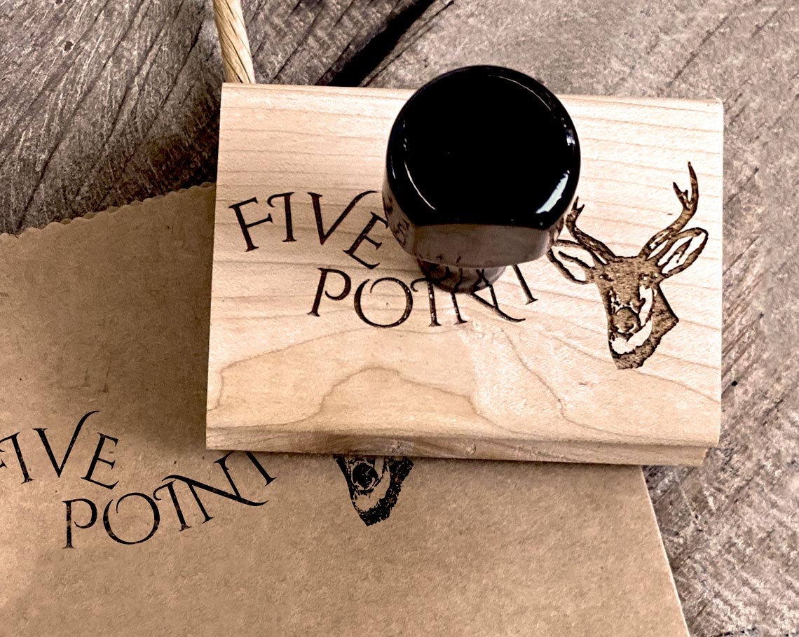 A custom logo rubber stamp with a deer head design and the text FIVE POINT stamped on craft paper next to the wooden engraved stamp tool on a rustic wood surface