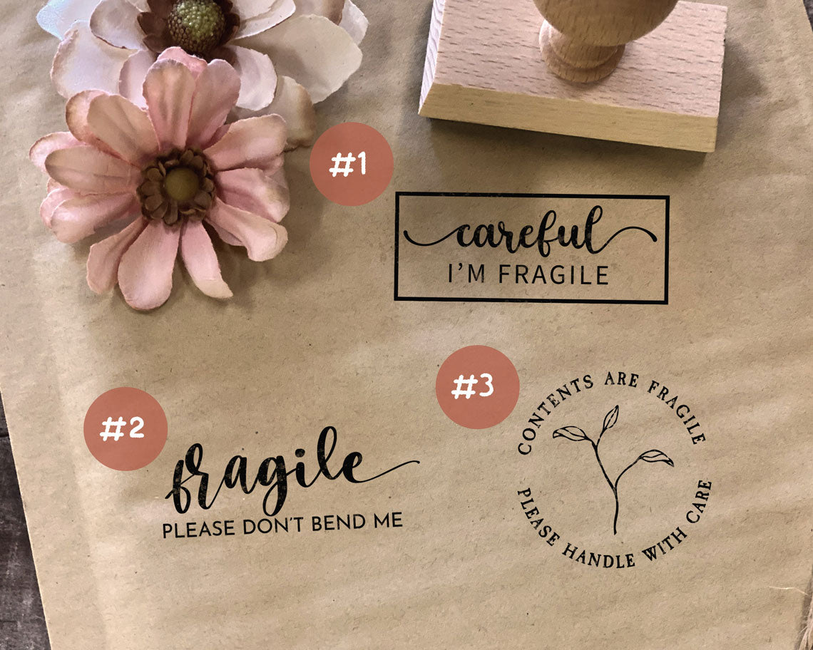 A craft paper with different fragile handling stamps including Careful Im Fragile and Please dont bend me along with a wooden stamp handle and decorative flowers on a wooden surface