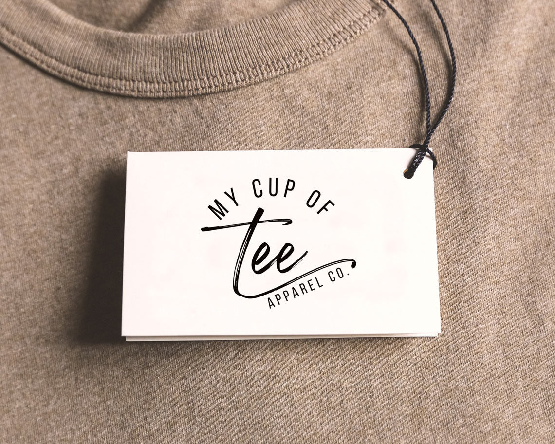 A clothing tag with My Cup of Tee Apparel Co logo printed on it attached to a grey shirt with a black string