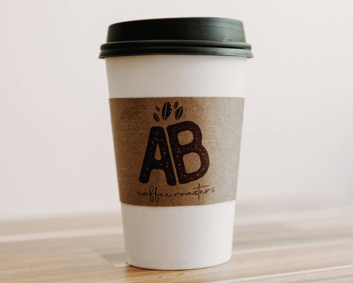 A disposable coffee cup with a craft sleeve stamped with the initials AB and the text coffee roasters along with a leaf design