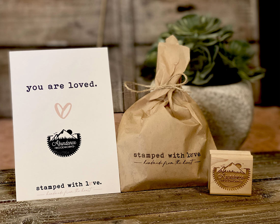 A customer thank you card stamped with the message you are loved and a laser engraved custom rubber stamp with a wrapped package in the background on a wooden table